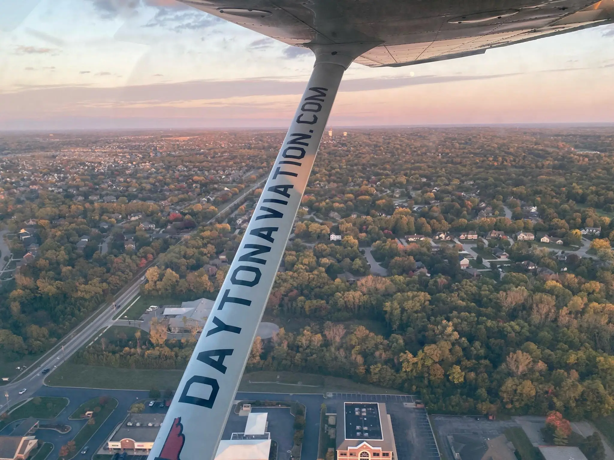 Airplane flying over city during the day seen from a cockpit window with visible right wings inscription of daytonaviation.com